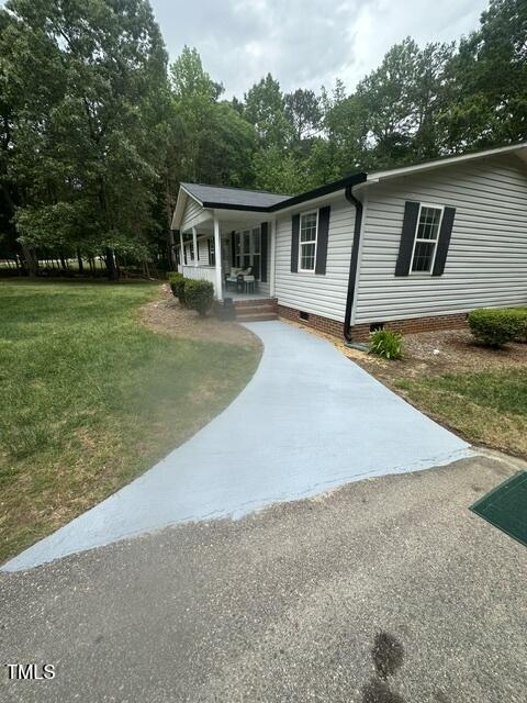 A light-colored concrete walkway leads to the front porch of a white house with black shutters, surrounded by grass, shrubs, and tall trees in the background.
