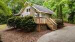 A small tan cottage with a stone chimney and wooden porch sits surrounded by trees and bushes. A short staircase leads to the raised entryway, and a concrete path curves in front of the house.