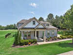 A modern two-story house with gray siding, white trim, and a covered front porch. The home is surrounded by green lawn, colorful flowers, and trees, with a driveway and a golf course in the background under a clear sky.