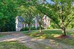 A two-story brick and siding house with white trim, surrounded by green trees and a grassy yard, sits on a spacious lot with a dirt driveway in front. Sunlight filters through the branches, creating a peaceful atmosphere.