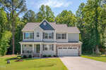 Two-story suburban house with a mix of gray siding and stone accents, attached two-car garage, front porch, and neatly landscaped lawn, surrounded by tall trees under a sunny blue sky.