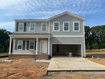 Two-story gray house with white trim, a covered front porch, and an attached two-car garage. The driveway and yard are unfinished, with dirt and construction materials visible around the property.