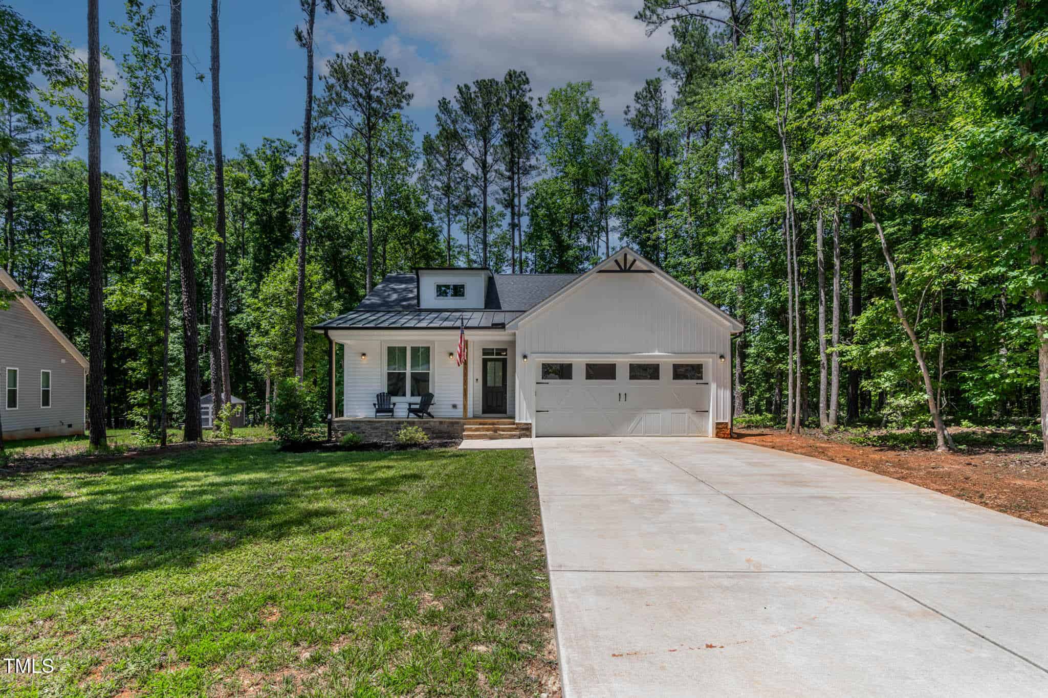 A modern white single-story house with a gray roof, double garage, small front porch with chairs, and a concrete driveway, surrounded by tall trees and green grass under a blue sky.