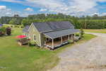 A green house with a front porch and solar panels on the roof sits on a spacious grassy lot, with a deck and red umbrella on one side and surrounded by trees under a partly cloudy sky.