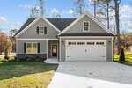 Single-family house with gray siding, white trim, and a double garage. Dark shutters frame the windows, and the home is surrounded by a neat lawn, young trees, and a driveway leading up to the garage.