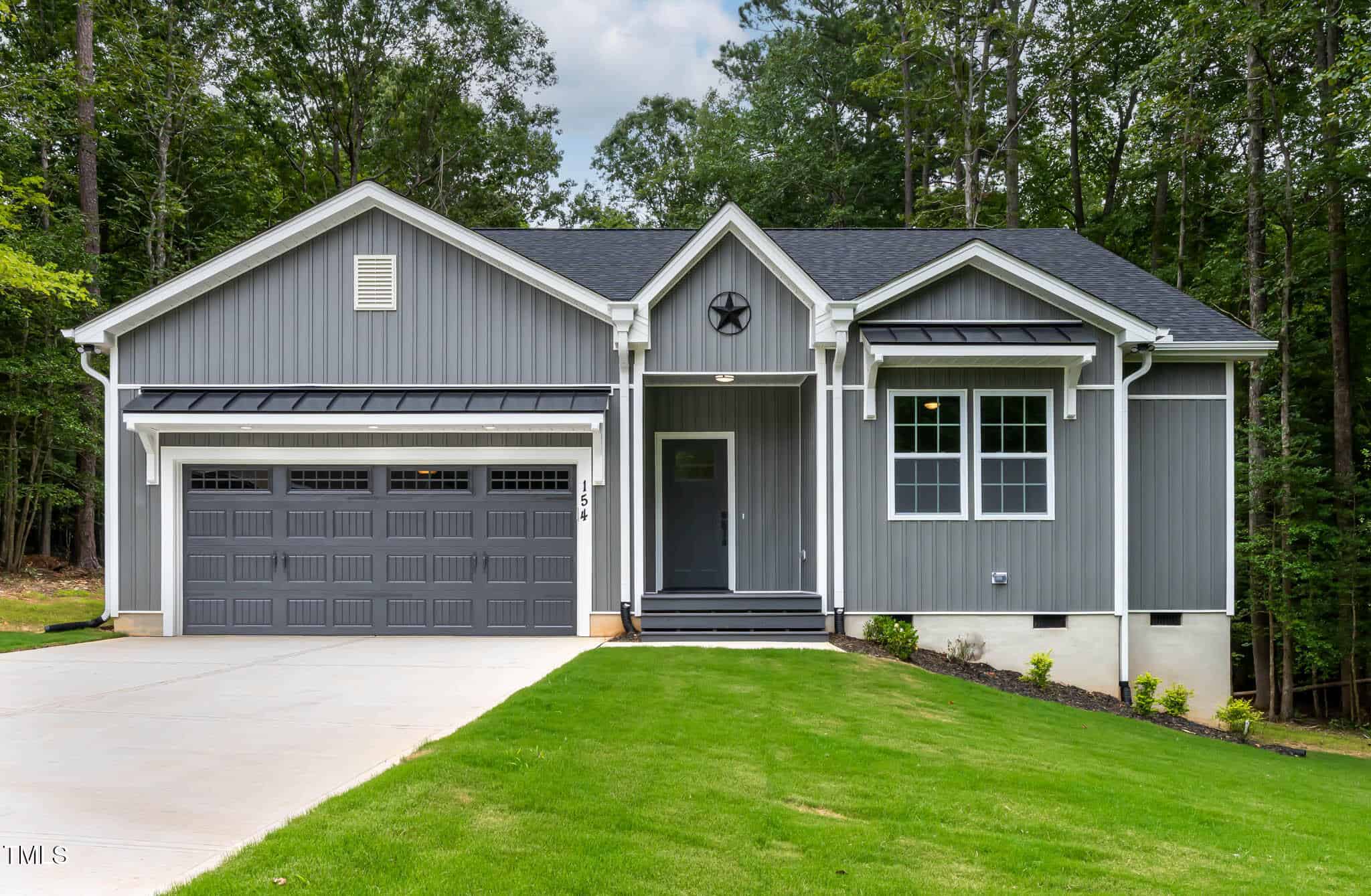 A modern, single-story gray house with white trim, a two-car garage, front porch, black door, and manicured lawn, surrounded by tall green trees.