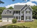 A two-story gray house with white trim and black shutters, featuring a double garage, front porch with steps, and surrounded by a green lawn and trees under a partly cloudy sky.