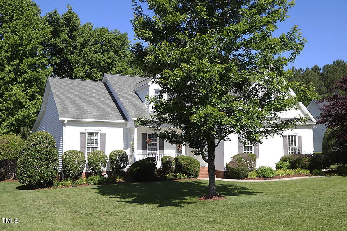 A white, single-story house with gray roof shingles, surrounded by neatly trimmed bushes and a well-kept lawn. A leafy green tree stands in the front yard under a clear blue sky.