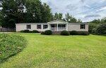 Single-story beige manufactured home with a white-railed front porch, brick foundation, and several bushes in front, set on a large, well-maintained grassy lawn with trees and shrubs in the background.