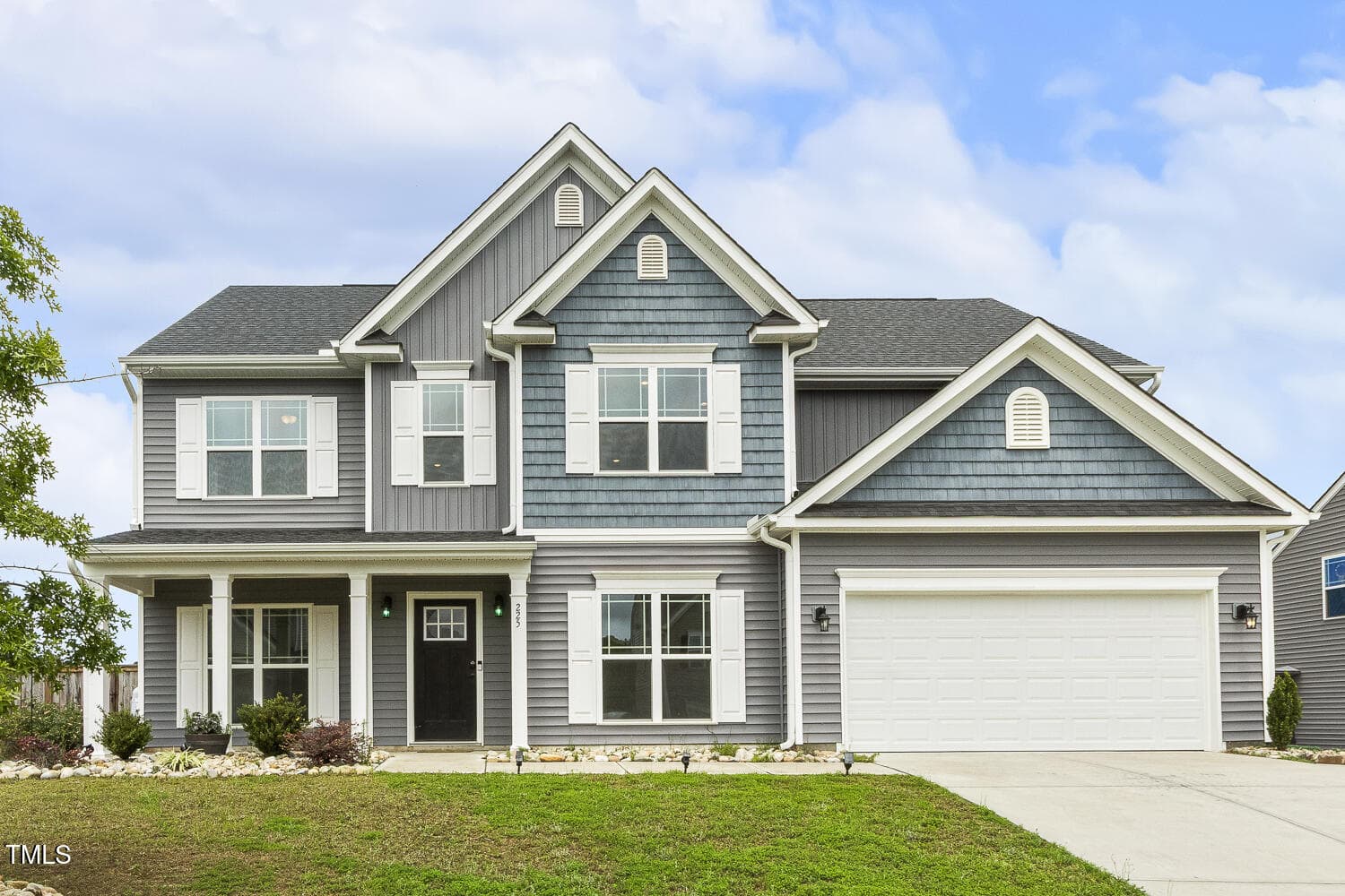 Two-story modern house with gray siding, white trim, a covered front porch, and a large attached two-car garage. The lawn is well-kept, and the sky is partly cloudy.