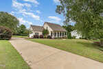 A beige house with a covered front porch, white columns, and brown roof sits at the end of a wide concrete driveway, surrounded by a well-maintained lawn, shrubs, and trees under a blue sky with scattered clouds.