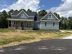 A newly built blue house with white trim, a covered front porch, and multiple gables sits on a yard with fresh straw and a paved driveway, surrounded by trees under a partly cloudy sky.