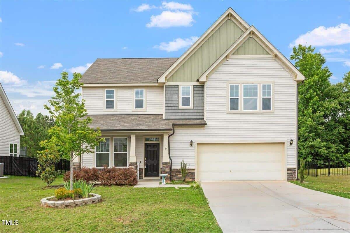 Two-story suburban house with light-colored siding, gabled roof, and attached double garage. There is a small front porch, a young tree, and a landscaped flower bed in the front yard. The sky is blue with scattered clouds.