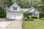 A light gray single-story house with white trim, double garage, arched window, and neatly trimmed bushes in front, surrounded by tall pine trees and a well-maintained lawn.