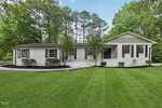 A single-story white brick house with black shutters and trim, set on a well-manicured green lawn, surrounded by tall trees and landscaping with bushes in front. Steps lead to a covered front entry.
