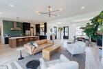 A modern open-concept living space with a tan leather sofa, light armchairs, a wooden coffee table, and a kitchen with green tile backsplash, barstools, and pendant lights. Large windows provide natural light.