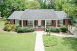 A single-story red brick house with a gray shingle roof, white trim, front porch, bushes along the front, a curved sidewalk, and a wooden accessibility ramp on the right side, surrounded by green lawn and trees.