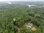 Aerial view of a wooded area with a house icon marking a specific spot among the trees; nearby are houses, roads, and a large body of water in the background.