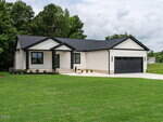 Single-story modern house with cream siding, black trim, a black roof, and a two-car garage. There are minimal landscaping features and the home is surrounded by a green lawn and trees in the background.