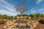A rocky fountain with flowers and a small pond sits in front of a large tree and three flagpoles flying American and other flags, surrounded by landscaped greenery and under a blue sky with wispy clouds.