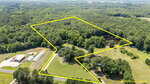 Aerial view of rural land with property lines outlined in yellow, showing fields, greenhouses, a pond, wooded areas, a house, outbuildings, and a road in the foreground. Text notes property lines as estimates.