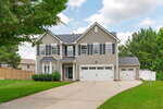 Two-story beige suburban house with white trim, black shutters, and a three-car garage. The house has a large driveway, green lawn, and is surrounded by trees and a wooden fence under a partly cloudy sky.