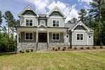 A two-story modern farmhouse with gray siding, white trim, stone foundation, double front doors, and a covered porch, set on a grassy yard with young shrubs and trees in the background.