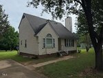 A small beige house with a steep roof, chimney, and front porch sits on a grassy lawn with large trees nearby. A driveway runs alongside the house. The sky is overcast.