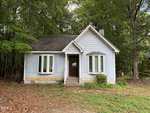 A small, light blue house with white trim and a dark roof stands in front of tall trees. The house has a central front door, two bay windows, and a patchy yard with sparse grass and fallen leaves.