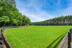 A wide, green grassy field bordered by dark wooden fences, with a row of trees on the right and left sides under a blue sky with light clouds. The sun casts bright light, creating a peaceful rural scene.