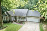 A gray, single-story house with a covered front porch, white trim, and an attached two-car garage. The home is surrounded by trees and has a neat lawn and a concrete driveway.