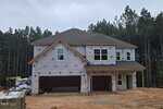 Two-story house under construction, wrapped in weatherproofing material with exposed wooden beams. Windows and garage doors are installed. Surrounded by dirt and construction materials, with trees in the background.