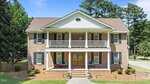 Two-story brick house with white columns, a covered front porch and balcony, wooden double front doors, and black shutters. The house is surrounded by green lawns, trees, and a driveway.