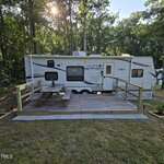 A white travel trailer is parked in a wooded area. In front, there is a wooden deck with a picnic table and simple railings. Sunlight filters through the trees in the background.