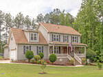 A two-story suburban house with gray siding, stone accents, and a covered front porch. The house has a manicured lawn, small shrubs, a U.S. flag, and is surrounded by tall trees.