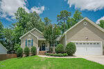A tan, single-story house with white trim, a black front door, and a two-car garage. Well-maintained shrubs line the front, and a lush green lawn leads to the entrance under a bright blue sky.