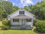 A white, single-story house with black shutters, a covered front porch, and metal railings. The lawn is green and well-kept, bordered by bushes, with trees in the background under a partly cloudy sky.