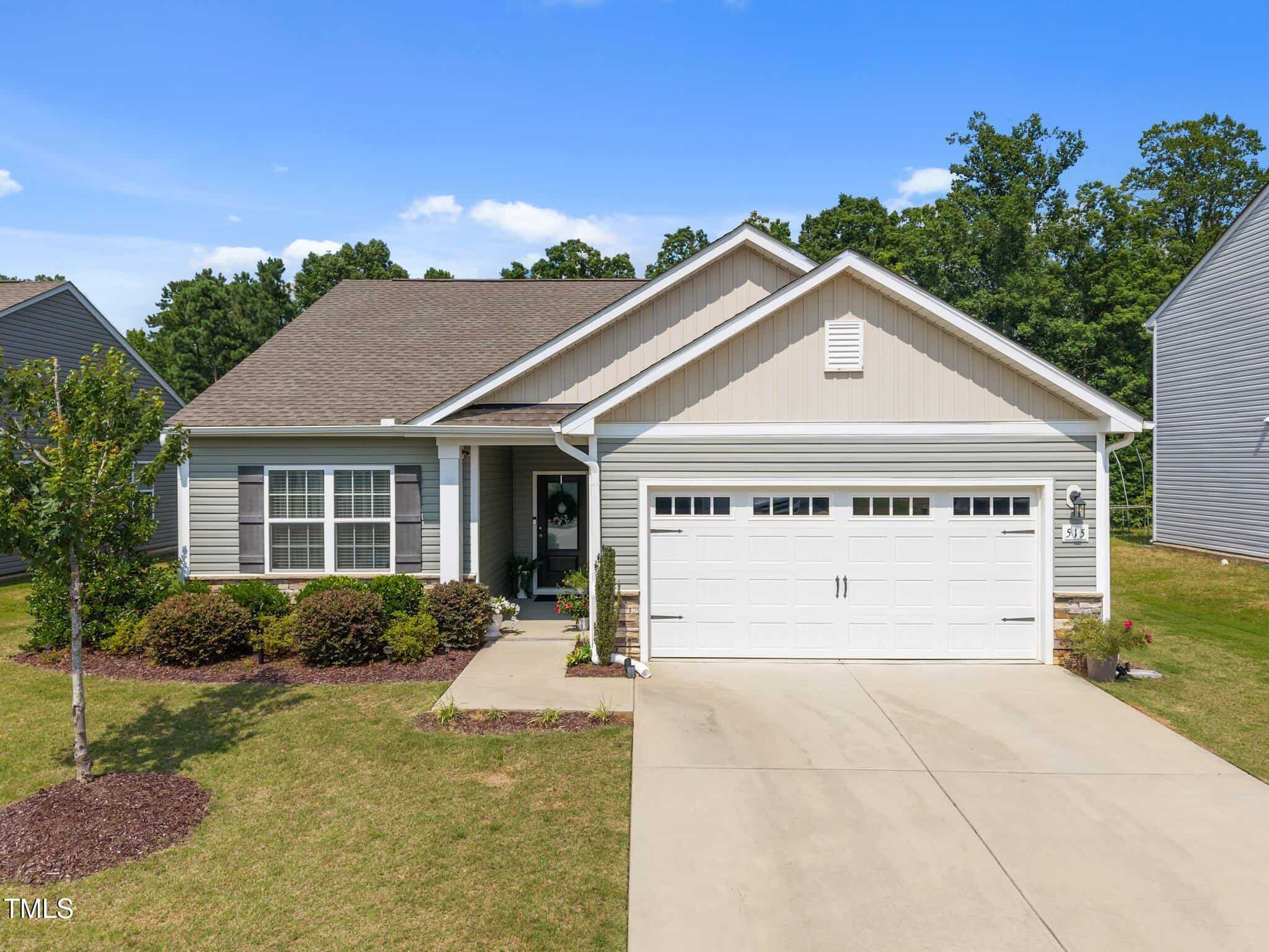 Single-story house with beige siding and white trim, two-car garage, small front porch with decorative wreath, neatly landscaped front yard with shrubs and a young tree, and a concrete driveway.