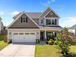 Two-story suburban house with a double garage, beige siding, dark brown gable accents, white trim, and a neatly landscaped front yard under a clear blue sky.
