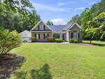 A single-story brick house with white trim and arched windows sits surrounded by green grass and trees under a blue sky with scattered clouds. A detached white garage is visible on the left.