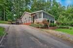 A light-colored manufactured home with a screened porch sits on a sloped driveway surrounded by lush green trees and landscaped bushes. The yard is well-maintained with a mix of grass, stone, and mulch beds.