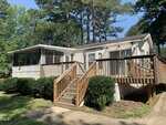 A white manufactured home with a screened porch, large sliding glass doors, and a wooden ramp and stairs. The home is surrounded by green shrubs, grass, trees, and is set in a wooded area on a sunny day.
