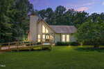 A single-story house with a peaked roof and large windows glows warmly at dusk. Surrounded by lush green lawn and trees, the house features a wooden ramp leading to the front entrance.
