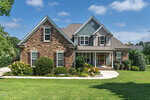 A two-story suburban house with a brick and gray siding exterior, white trim, and gabled roof, surrounded by green lawn, shrubs, and trees under a blue sky with clouds.
