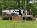 A white camper trailer sits behind a large wooden deck with stairs, black railings, patio furniture, potted flowers, and an American flag, surrounded by green grass and tall trees.