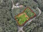Aerial view of a wooded residential lot outlined in red, located at the intersection of two roads and surrounded by trees, with a nearby house visible at the top of the image.