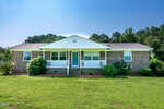Single-story brick house with white trim and a covered front porch, surrounded by green shrubs and grass, set against a backdrop of trees under a clear blue sky.