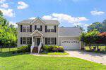 Two-story beige house with black shutters, white trim, and a front porch. It has a two-car garage, a paved driveway, green lawn, shrubs, and trees. The sky is blue with scattered clouds.