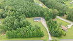 Aerial view of a house with a gray roof surrounded by dense green trees, a gravel driveway, and a few parked cars; bordered by a main road and neighboring buildings in a rural area.