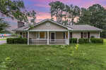Single-story house with a front porch, light-colored siding, and a gray roof, surrounded by green grass and trees under a pink and purple sunset sky.